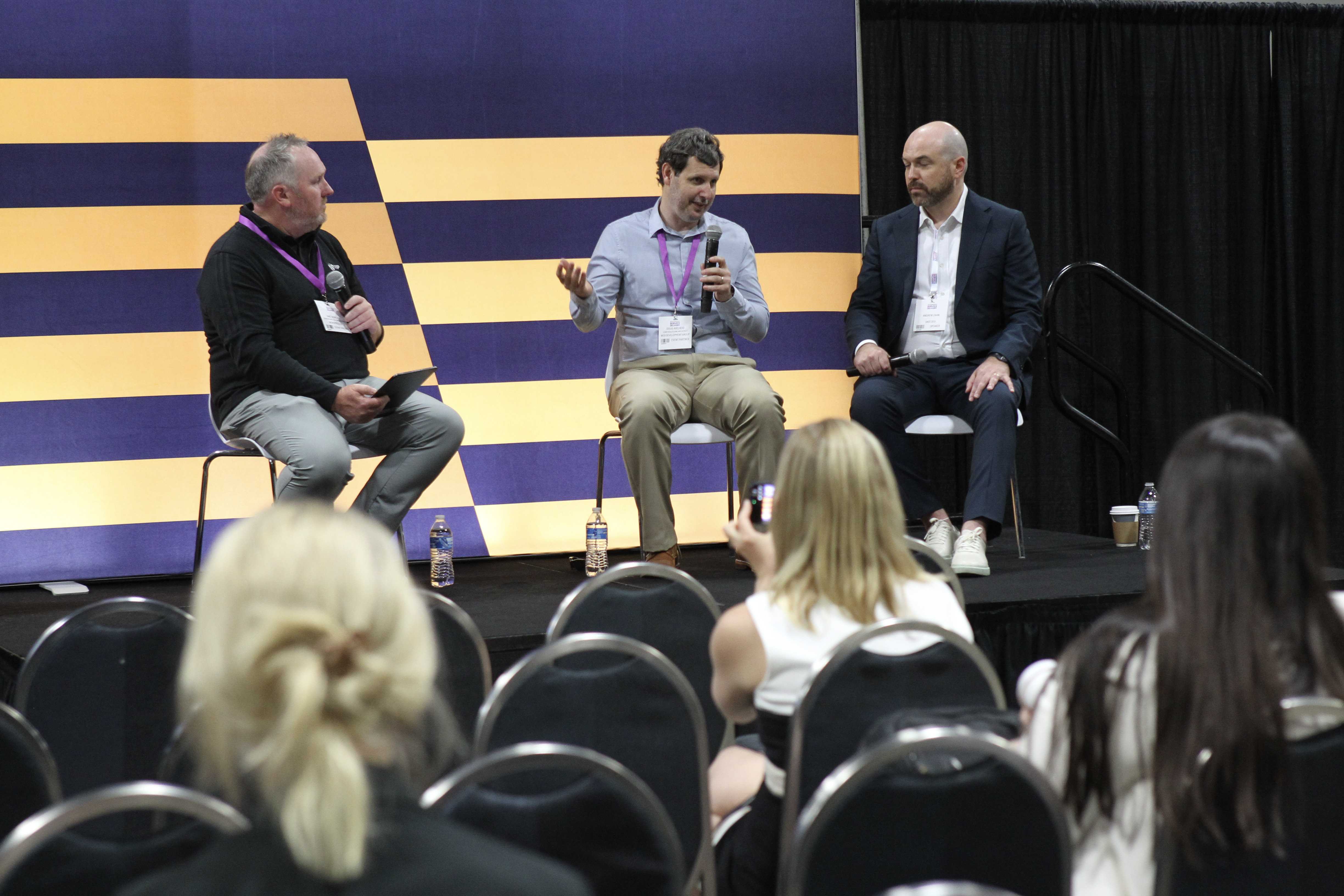 Jake Spurlock, Doug Axelrod, and Andrew Zahn seated on stage during a panel discussion at the Government Service Delivery 2026 conference in Washington, DC. Doug, in the center, speaks into a microphone and gestures with one hand while the other two panelists listen. Behind them is a purple and gold striped backdrop, and audience members are seated in the foreground.